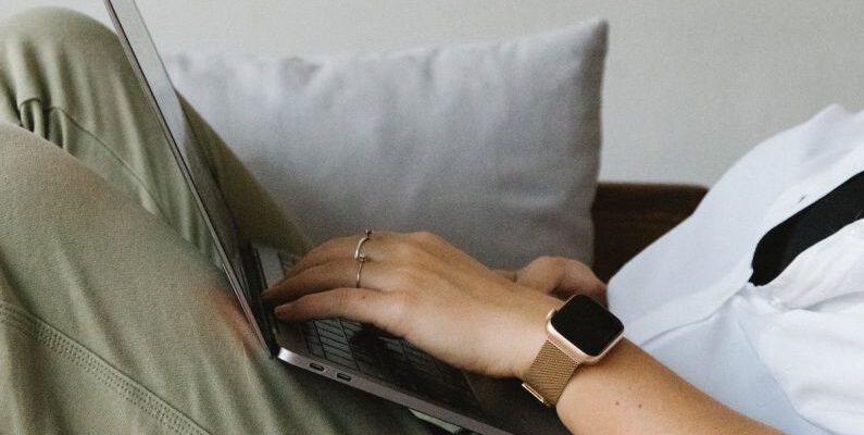 Remote Work Challenges - Side view of crop unrecognizable female freelancer in casual clothes and smartwatch lying on comfortable sofa and working remotely on laptop